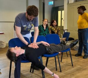 A man acts out performing heart compressions on another man stretched out on chairs, apparently unconscious, while others look on
