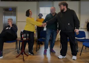 Three actors shake hands as though doing a deal, while another looks on and one sits at the side having a drink and appearing disinterested