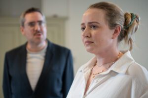 A woman stands stony-faced, with a man looking on