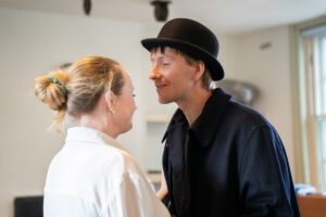 A man in a bowler hat stands smiling over a woman up close