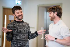 Actors in rehearsal - one is speaking animatedly, while theatrically gesturing with a champagne flute in hand, while the other looks on and smiles.