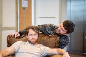 Two actors in rehearsal, one apparently teasing another from behind the chair they're sitting in.