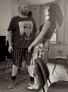 A male and a female actor stand facing each other smiling