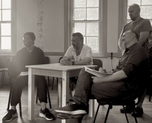 Three men sit at a table and one stands, looking on at a scene in rehearsal