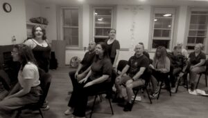 Image of several actors sitting on chairs as though on a plane, and a couple of others standing alongside.
