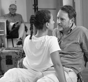 A male and female actor sit entwined on the floor looking into each other's eyes, with a man sitting in the background looking on