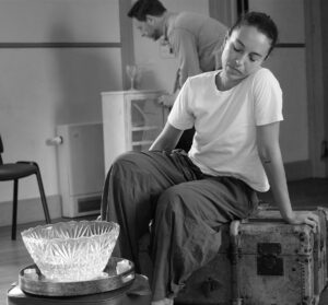 A female actor sits on an old chest looking at a glass bowl before her, with a male actor pouring a drink in the background