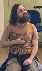 A heavily-bearded maeleactor kneels on the floor, looking up with a smirk on his face