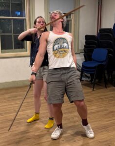 A female actor in a threatening pose stand behind a male actor and holds her sword to his throat