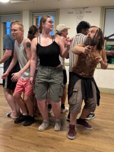 A group of actors stand in a circle with their backs to each other, looking out for danger