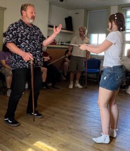 A male actor stands appealing to a female actor who has drawn her sword on him