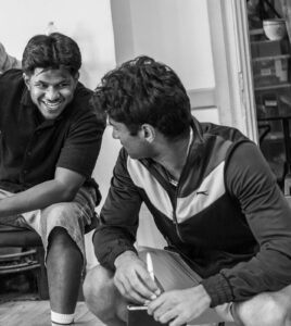 A male actor leans forward in a chair with a broad smile, while crouching at his feet looks over his shoulder at him