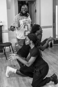 A male and a female actor kneel alongside each other miming typing on a keyboard, while a second male actor stands behind them looking fierce with a clenched fist