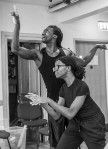 A male actor stands with his arms outstretched in an elegant pose pointing upwards, while a female actor crouches in front of him with her hands outstretched as though typing