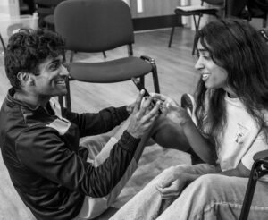 A male and a female actor sit close together on the floor, touching hands and smiling