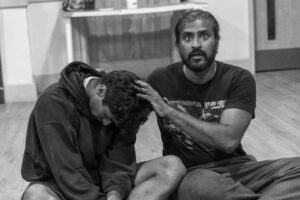 Two actors sit cross legged on the floor, one with his hands on the bowed head of the other
