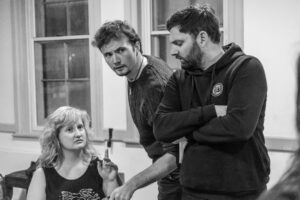 A female actor sits at a table looking left with her finger pointing upwards, while one male actor stands looking down at her and another stands beside her also looking to his left.