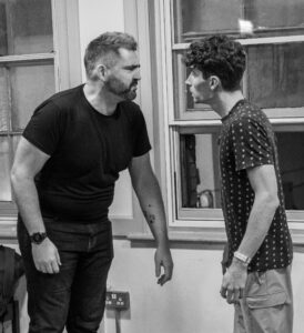 Two male actos stand head to head in close conversation
