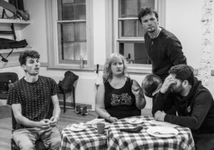 Two male and one female actors look to their left in shock at someone off camera, while a third male actor sits at the same table with his hand covering his face