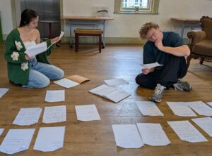 A male and a female actor sit on a floor surrounded by and reading papers