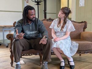 A male and a female actor sit in conversation on a sofa
