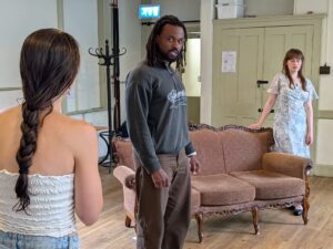 A male actor stands between two female actors and looks accusingly at the one with her back to the camera