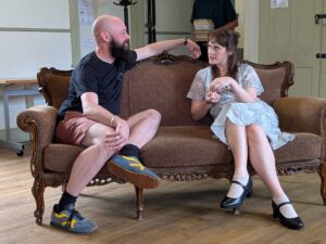 A male and a female actor sit facing each other and talking on a sofa