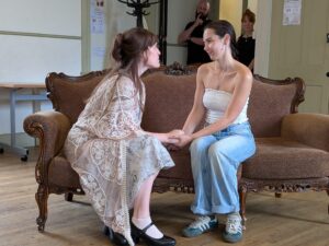 Two female actors sit on a sofa in earnest conversation, holding hands