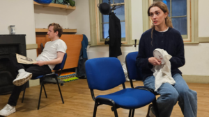 Aman holding a newspaper and a woman clutching a piece of embroidery sit in chairs looking away from each other