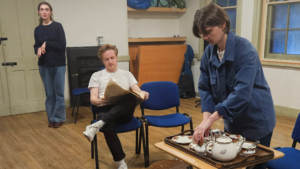 A man sits reading a newspaper between two women, one of them standing looking into the distance, the other, setting out a tea set on a tray