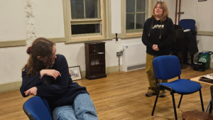 A woman in distress sits in a chair, while another stands looking towards her with concern.