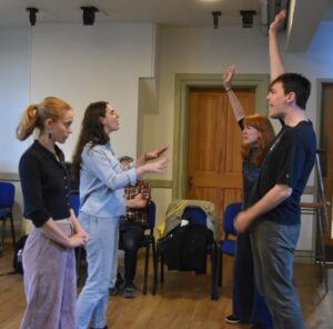 Three female and one male actor stand facing each other, two of them with an arm raised