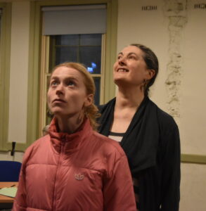 Two female actors, one behing the other, stand looking upwards to the sky.