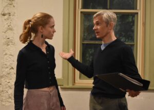A male and a female actor stand in conversation, he with a file in his hand and explaining a situation